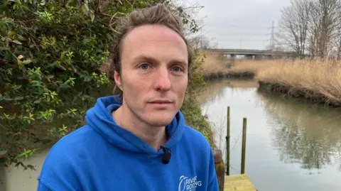 Paul Powlesland from the River Roding Trust. He is a man with light brown hair pulled behind his head and is wearing a blue hoodie, sitting by the edge of a river on the right and then in the background there is a flyover across the river, with telephone lines.