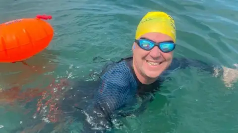 Edward Sault treading water in the sea in a yellow swimming hat and blue reflective goggles with a orange swim buoy