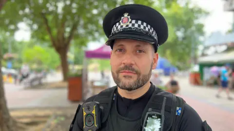 A policeman with a short beard looking at the camera with a treelined street out of focus behind him.