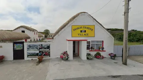 Google A white thatched building with a yellow sign above the door reading "Doagh Famine Village". There is a concrete path outside the building and an electricity pole to the right of it. There is a cloudy sky in the background.