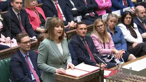 Chancellor Rachel Reeves standing at the dispatch box in he House of Commons reading from a folder, with fellow MPs behind her on green benches