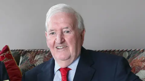 Lady Bloomfield has short brown hair and is wearing a white shirt with a navy cardigan. Sir Kenneth Bloomfield has short grey hair and is wearing a navy suit blazer, white patterned shirt and a red tie.