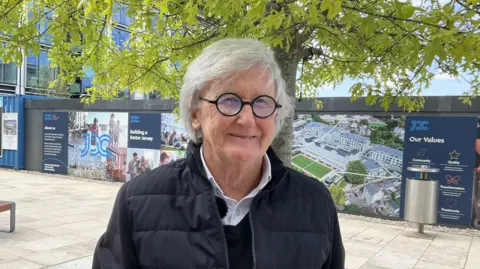Joe Dickinson pictured smiling at the waterfront business district. He has white hair and round glasses. He's wearing a navy blue jacket and black jumper.
