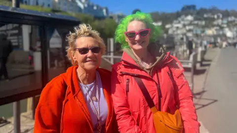 Two women stood side by side, both are wearing sunglasses and smiling. The woman on the left has short blonde hair and is wearing a purple top and red zip up hoodie. The woman on the right is wearing a red coat and has an orange bag and has short green hair. Behind them is a seaside town. 