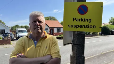 An image of Neil Campbell who is wearing a yellow polo top, he has his arms folded and is standing to the left of a yellow sign with the words Parking Suspension on it.