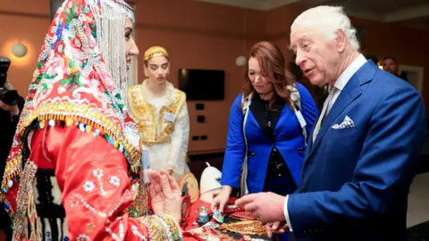 King Charles speaks to a woman in wearing an elaborate embroidered headscarf and dress. The King is wearing a blue suit with a light coloured tie. 