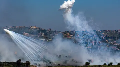 Two explosions of white smoke in front of a hillside - the Lebanese side of the border with Israel