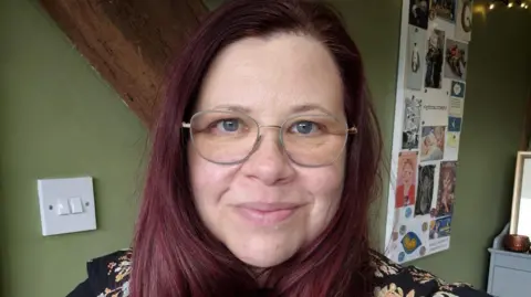 The image shows a woman standing indoors against a green wall. The woman has long, wavy, dark reddish‑purple hair that reaches past the shoulders. They are wearing a dark top with a floral or patterned design in lighter colours, along with a necklace that has a small, circular pendant