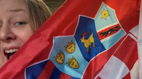 Emma Gayle with her mouth wide in an excited expression, holding up a Croatian flag which is partly obscuring her face