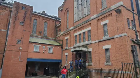 A large, L-shaped red brick structure. Part of the building is shaped like the entrance to a chapel, and has large windows at the top. There are doors where five or so people are stood on steps.