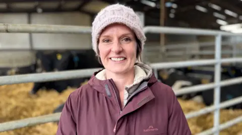 Dallas New a woman with short, brown hair, smiles at the camera while standing in a large cattle shed. She is wearing a light pink wooly hat and a plum-coloured hooded coat. There are number of cows behind a metal gate in the background.