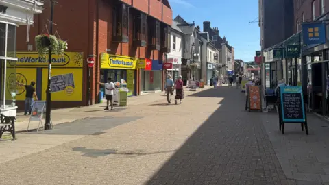 A high street on a sunny day. The street is pedestrianised and lined with shops. It is a sunny and a few people are walking.