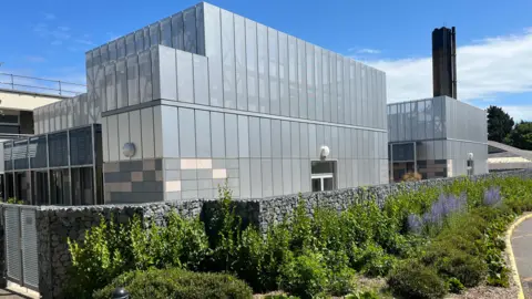 BBC A large grey building, behind some flowers. The Clinical Care Unit at the Princess Elizabeth Hospital. It has a big black chimney rising above it. 