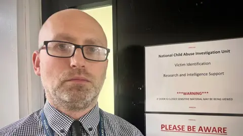 Detective Smith - a bald man with glasses and a beard, looks straight into the camera inside a police station. Behind him on a door, we see a printed sign which reads: "Warning - if door is closed sensitive material may be being viewed."