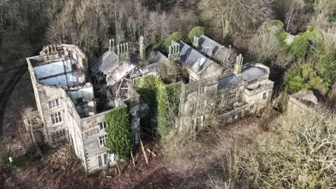 Photograph of Winstanley Hall in Wigan. The image shows an overgrown, derelict building, with collapsed walls and no roof.