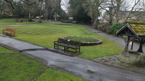 View of a park with large areas of grass and patches of freshly laid turf. A well and two benches stand alongside a concrete path. There are trees and buildings in the background.