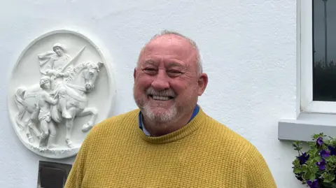 A man is pictured in a yellow cable knit jumper standing outside St Martin parish hall. He is grinning. He has short grey hair and stubble.