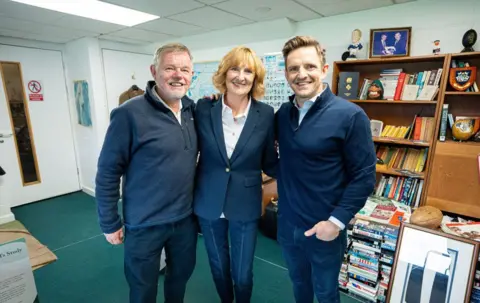 Scottish Rugby A woman in a dark blue jacket and white blouse with reddish blonde hair stands between a man with greying hair and a beard in a blue zip up top and a younger man with spiky blond hair in a blue top and jeans. They are in a recreation of Bill McLaren's old research room
