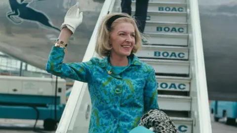 Fox Photos / Getty The woman from the above pictures, this time in colour. She is descending the steps of a BOAC aircraft and smiling while waving a white-gloved hand. She is wearing a blue and green suit and is carrying a leopard print coat over her arm. Her blonde hair is pushed back with a black band.