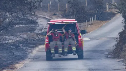 SFRS Two fire officers, one female, are in the back of a red fire vehicle with the boot door open. They are wearing fire safety clothing and around their necks are breathing masks. Both wear sunglasses. Around them are the remnants of burnt trees and grassland. The red vehicle appears to be driving on a road away from the camera.