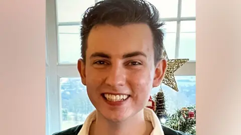Tracy Scott Ethan, who has short brown hair, looking directly at the camera and smiling. There is a window and a Christmas tree behind him.