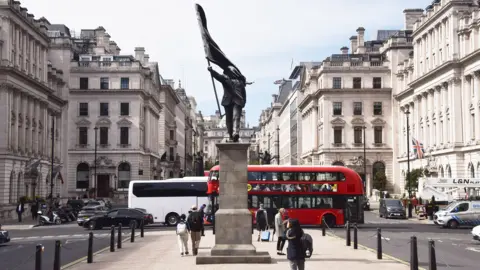 A grey statue of a man wearing a suit, walking forward off a plinth, carrying a flag that covers his face. A red London bus can be seen in th bcakground