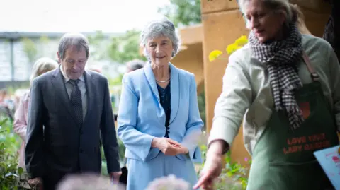 PA Media A woman in a blue suit and blue dress, with a navy top looks down at an out-of-focus flower bed