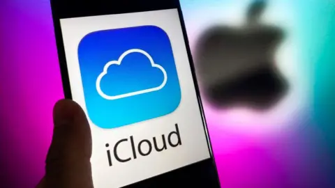Getty Images A phone with the words iCloud in black letter, and the iCloud blue and white logo of a cloud on it, against a white background. A blurred Apple logo in black is behind it against a purple background.