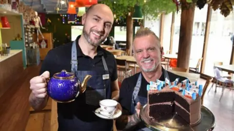 Richard Wilson (R) and Neil Senior (L) who both own Cake'ole in Skipton, stood inside their shop holding teacups and cups and a chocolate cake.