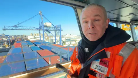 Man in reflective orange safety gear stands on the bridge of a cargo ship in the Port of Immingham. A window behind him overlooks the deck of the ship, where blue container boxes are being loaded using a crane out.