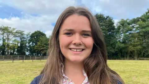 A woman looking directly into the camera and smiling. She's wearing a blue knitted jumper with a pink checked shirt underneath, and has long brown hair. 