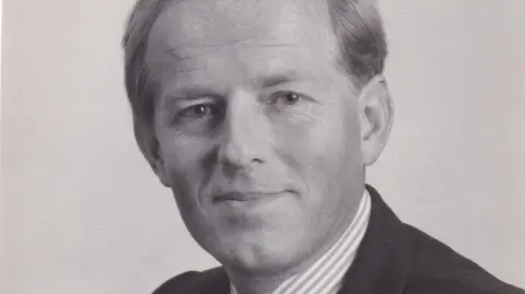 Passion Pictures A black and white professional portrait of John Smyth wearing a stripey collared shirt and dark suit. He is smiling at the camera and appears friendly. 