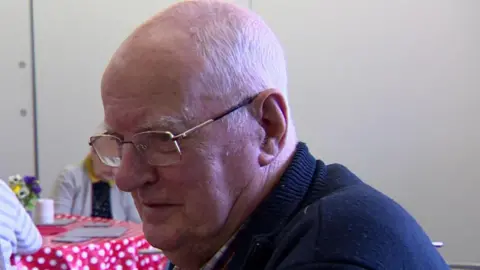 Eddie - an elderly man with short, white hair - is pictured during a BBC interview. He is wearing metal framed classes and a navy jumper over a checked red, white and navy shirt. Two women are seated at table in the background. It is covered with a red and white spotted table cloth and a vase of flowers. 