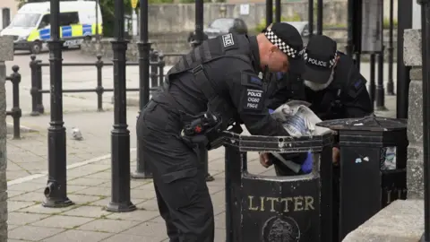 Two police officers are seen looking through a bin, with the Daily Mail newspaper in one officer's hand.