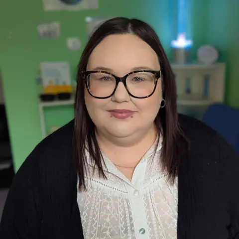 Frankie, a woman with medium-length dark hair and wearing glasses and a white lace blouse under a black cardigan, looks at the camera