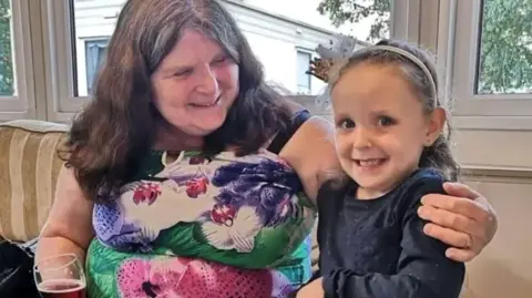 A woman with long brown hair and a flowery dressed smiles as she looks at and puts her arm around a young girl while sitting down and holding a glass of red wine in her right hand. The girl standing next to her has dark brown hair and is wearing a white headband and dark blue outfit. 
