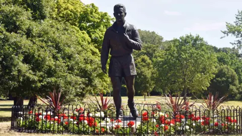 A life-size, grey sculpture of Brian Clough. He appears to be walking with his football boots slung over his shoulder by their laces. The figure has short hair and is wearing shorts and a long sleeve top. The statue is surrounded by red and white flowers and there are trees in the background.