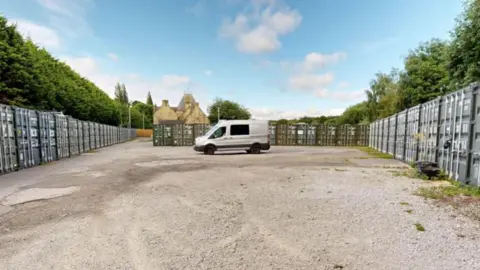 A storage yard, with shipping containers around the outside and a large concrete forecourt. A silver van in parked in the centre.