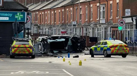 Leicester Media Online Two police cars at a junction. Two damaged cars, one flipped on its side can be seen, with debris in the road