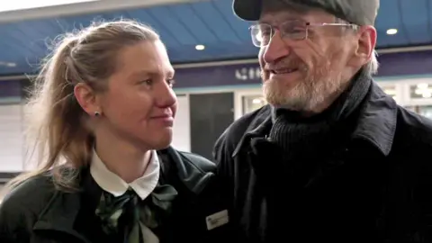 Veronika Rogers, who has brown hair in a ponytail and wears a GWR uniform, smiles at Ian Drewery, who has a grey beard, a flat cap and glasses.