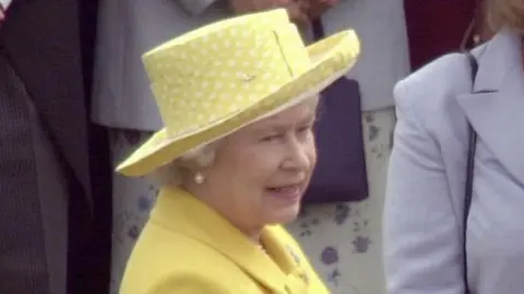 The late Elizabeth II attending a garden party at Buckingham Palace in July 2000. She is wearing a bright yellow coat dress, a matching hat and pearl earrings.