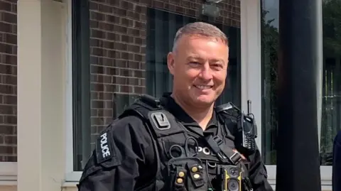 PC Scott Caswell with short grey hair stands in his full police uniform in front of windows of a building behind.