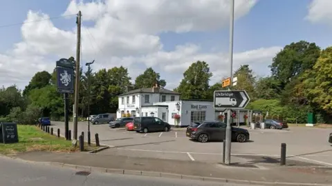 Google A roadside scene shows a white, two‑storey pub called the “Red Lion” with a small car park in front, where several vehicles are parked. A black pub sign mounted on a tall post stands to the left, and a road sign in the foreground points towards Uxbridge. The pub is surrounded by greenery and trees, with a grassy verge and pavement along the road. Overhead, a bright blue sky with scattered white clouds stretches above the scene, and lampposts and utility poles line the roadside.