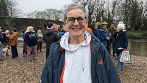 Sarah Preece, Cornwall councillor for Lostwithiel and Lanreath, on the banks of the River Fowey smiling at the camera. There are people gathered behind her.