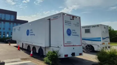 Clatterbridge Centre A large white trailer sits in a car park, with blue signage reading 'Lung Cancer Screening'