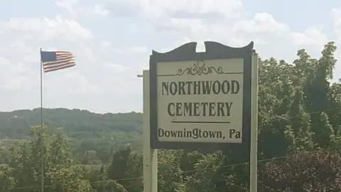 Duffy's Cut Project A sign reads Northwood Cemetery, Downington, Pa. Trees and wooded-hills are in the background and an American flag flies beside it