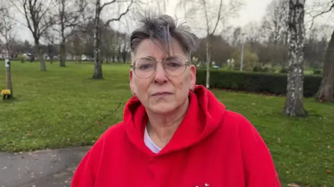 BBC The photo shows a woman looking at the camera. She has a straight face, and short grey hair. She's wearing a bright red hoodie. She's standing in front of a park, which has grass and trees in the distance. 