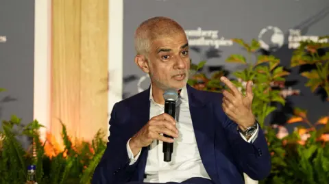 PA Media Khan sitting in a panel holding a microphone wearing a dark blue blazer and white shirt.