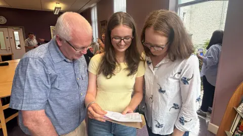 Dad Killian is to the left with grey hair, wearing a blue shirt and light trousers. Aideen is in the middle wearing a yellow top and denim jeans. She has brown hair and glasses. She is holding her results. Her mother Laura is to her right, with shoulder length brown hair with glasses.