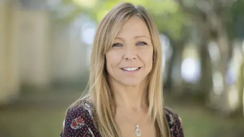 Claire Chick with long dark blonde hair. She has a silver necklace and a dark blue and red patterned top.